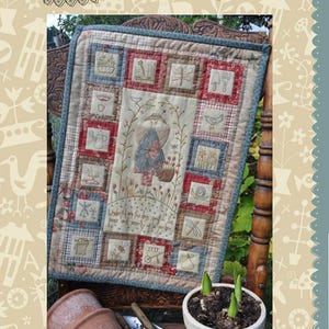 May include: A quilted wall hanging with a patchwork design featuring a variety of embroidered images, including a woman in a garden, birds, flowers, and a watering can. The text "Underneath are the days spent in the peace and serenity of your garden" is embroidered on the wall hanging. The wall hanging is displayed on a wooden chair with a potted plant in front of it.