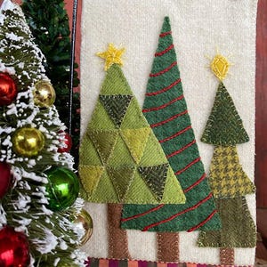 May include: A white felt wall hanging with three green felt Christmas trees. The trees are decorated with red and yellow felt details. The bottom of the hanging has a brown felt border with a plaid ribbon.