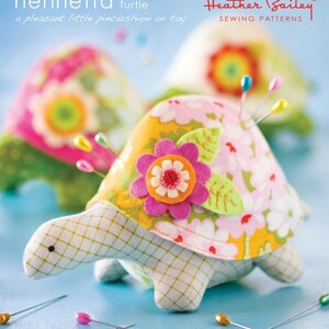 May include: A handmade turtle pincushion with a floral design on its shell. The turtle's shell is decorated with pink and white flowers, a large pink flower, and green leaves. The pincushion is surrounded by colorful pins. The text "Henrietta Turtle" is visible.