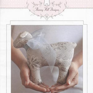 May include: A gray and white fabric lamb with a white satin bow, held in the hands of a person. The lamb is labeled "Lollipop".