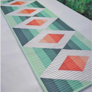 May include: A rectangular quilted table runner featuring a geometric pattern of white diamonds with coral centers. The background consists of alternating diagonal stripes in shades of dark teal, forest green, and mint green, finished with a mint green border.