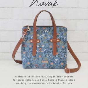 May include: A blue floral tote bag with brown leather handles and a shoulder strap. The bag has a zipper closure and metal hardware. Text on the bag reads "Novak" and "Sallie Tomato". The bag is designed for organization.