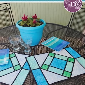 May include: Stained Glass Placemats pattern by Kris Holderness. The cover features two quilted placemats with blue and green geometric patterns, a blue pot with red flowers, and two glass cups. Dimensions are 18 inches by 12 inches.