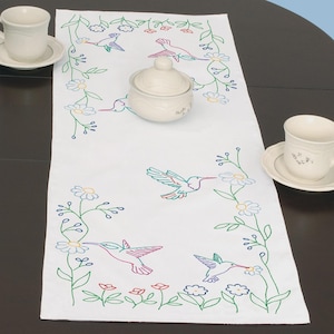 May include: White embroidered table runner featuring colorful hummingbirds and floral designs. The runner has a central design with a sugar bowl and two teacups on either side. The embroidery includes green, blue, red, and yellow thread.