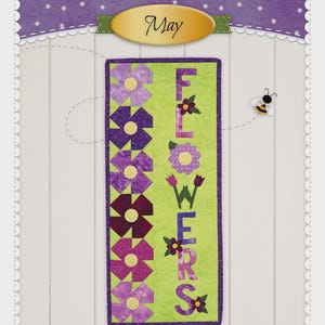 May include: A wall hanging with the text "A Year in Words" and "FLOWERS". The design features purple flowers and the word "FLOWERS" in purple letters on a green background. A small bee is also present.