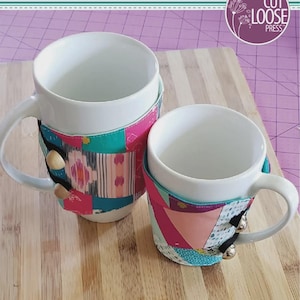 May include: Two white ceramic mugs wrapped in fabric cozies featuring a patchwork design of pink, teal, and patterned fabric scraps. Each cozy is secured with two round buttons and black elastic loops. The text reads Scrappy Mug Cozy, 5" x 14".