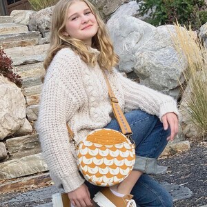 May include: A round, mustard-yellow and white patterned crossbody bag with a brown strap. The bag is labeled "ROUNDABOUT BAG #260" and features the text "Cotton Street Commons" at the top and bottom. The bag is part of a fashion accessory collection.