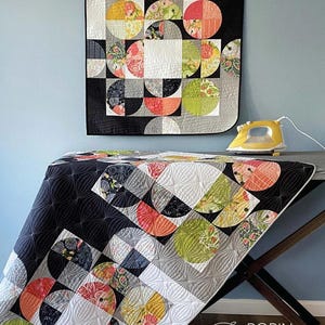 May include: A quilt pattern titled "Partial Eclipse" featuring a design of colorful half-circles on a black and white background. The quilt is displayed on a table with a yellow iron. The pattern is available in wall size 32.5 x 32.5 inches or large 64.5 x 64.5 inches.