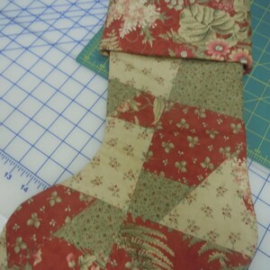 Op de afbeelding: Een gewatteerde kerstkous met een patchworkontwerp van rode, cr&egrave;mekleurige en saliegroene bloemenstoffen. De boord is gemaakt van bijpassende rode bloemenstof en aan de bovenhoek zit een klein stoffen lusje om de kous mee op te hangen.