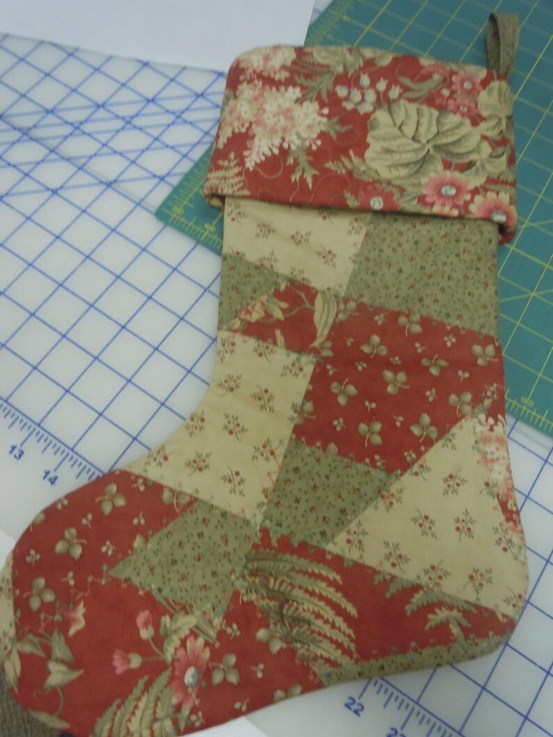Op de afbeelding: Een gewatteerde kerstkous met een patchworkontwerp van rode, cr&egrave;mekleurige en saliegroene bloemenstoffen. De boord is gemaakt van bijpassende rode bloemenstof en aan de bovenhoek zit een klein stoffen lusje om de kous mee op te hangen.
