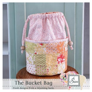 May include: A patchwork bucket bag with a drawstring closure. The bag features a mix of floral and patterned fabrics in shades of orange, yellow, and green. The top portion is a pink fabric with printed text. The bag is labeled "The Bucket Bag" with the tagline "Fresh designs from a Wyoming farm."