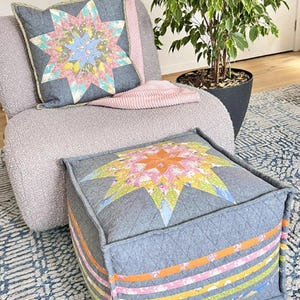 May include: A gray square ottoman and matching pillow with a colorful starburst design. The ottoman has horizontal stripes in shades of orange, pink, and green. The pillow rests on a gray chair with a pink blanket.