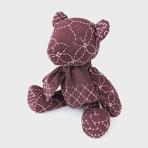 May include: A handmade burgundy teddy bear with white stitched floral patterns. The bear has a matching scarf and a button detail. The bear is sitting with its arms and legs extended.