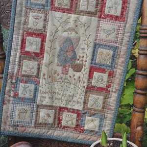 May include: A patchwork quilt with a blue and white floral design. The quilt is made up of squares with different designs, including a watering can, a bird, a dragonfly, and a snail. The quilt is hanging on a wooden chair. The text on the quilt reads: "Wonderful are the days spent in the peace and serenity of a garden."