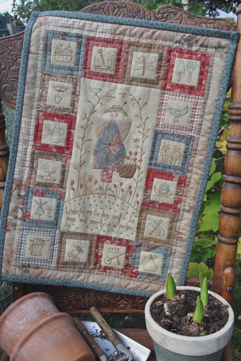 May include: A patchwork quilt with a blue and white floral design. The quilt is made up of squares with different designs, including a watering can, a bird, a dragonfly, and a snail. The quilt is hanging on a wooden chair. The text on the quilt reads: "Wonderful are the days spent in the peace and serenity of a garden."