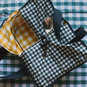 May include: A quilted casserole carrier featuring a black and white gingham exterior with black fabric handles. The interior is lined with yellow and white gingham fabric and contains a clear glass baking dish and a silver metal serving spoon.