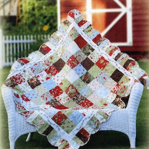 May include: A handmade quilt with a patchwork design in red, green, brown, and blue floral patterns. The quilt is draped over a white wicker chair. The edges of the quilt have a scalloped trim.