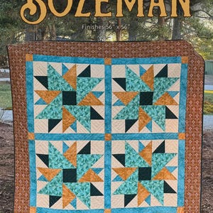 May include: A square quilt featuring four large star blocks in teal, mustard yellow, and dark green, set against a cream background. The quilt has a wide brown border with a small geometric pattern and measures 56 inches by 56 inches.