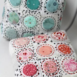 May include: Two square fabric pincushions featuring a grid of white squares with black dashed patterns and circular fabric appliqués. The top cushion has teal and green circles, while the bottom cushion features pink and orange patterned circles.