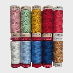 May include: A collection of Aurifil thread spools in various colors, including pink, green, yellow, red, blue, and brown. The spools are made in Italy and are labeled with the brand name and thread type. The thread is wound on wooden spools with red bases.
