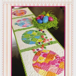 May include: A patchwork Easter egg table runner with colorful egg designs. The runner features a white background with green accents and a pink border. The image also includes a nest with Easter eggs and scattered jelly beans. The text "Shabby Fabrics" and "Patchwork Easter Egg Table Runner" are visible.