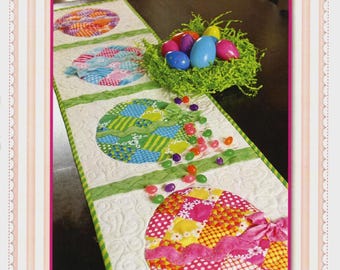 May include: A patchwork Easter egg table runner with colorful egg designs. The runner features a white background with green accents and a pink border. The image also includes a nest with Easter eggs and scattered jelly beans. The text "Shabby Fabrics" and "Patchwork Easter Egg Table Runner" are visible.