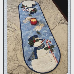 May include: A quilted, oval-shaped table runner with a winter theme. The runner features appliqued snowmen, snowflakes, and doves on a light blue background. A red candle sits in the center. The text "Winter Magic" and "Quilted Garden Designs" are visible.