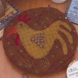 May include: A brown felt pincushion with a yellow felt chicken design. The chicken has a red comb and wattle, and a brown and white patterned wing. The pincushion is surrounded by green felt grass.