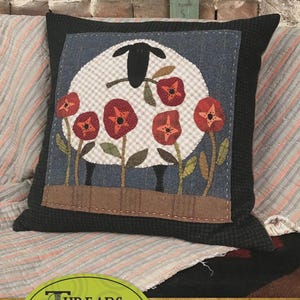 May include: A decorative pillow with a black border features a wool applique design. The design includes a sheep with a checkered body, red poppies, and the text "Threads that Bind". The pillow measures 14 x 14 inches.