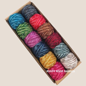 May include: A cardboard box filled with twelve colorful yarn balls. The yarn balls are in shades of blue, green, red, brown, pink, and gold. The text "RUSTY ROOF FABRICS" is printed on the bottom right corner of the box.