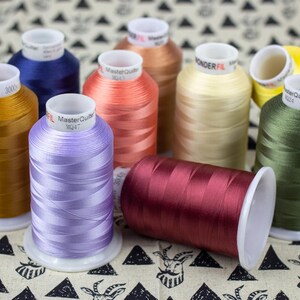 May include: A collection of spools of thread in various colors, including blue, orange, yellow, green, and purple. The spools are labeled with the brand name "MasterQuilter" and are arranged on a patterned surface. One spool is lying on its side.