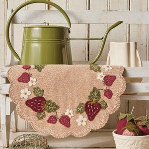 May include: A round, tan strawberry penny mat, 12 inches in diameter, with a scalloped edge. The mat is decorated with embroidered strawberries and white flowers. A green watering can and other decorative items are in the background.