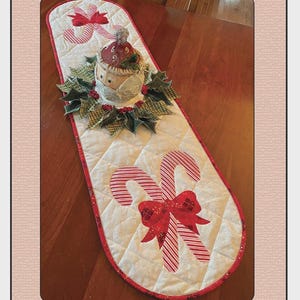 May include: A quilted Christmas table runner with candy cane and holly designs. The runner features red and white candy canes, red bows, and a decorative centerpiece. The text "Candy Canes & Holly" and "Quilted Garden Designs" are visible.