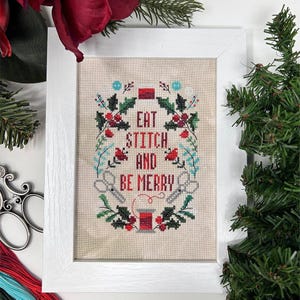 May include: A framed cross-stitch piece with the text "EAT STITCH AND BE MERRY" surrounded by a wreath of holly, berries, and sprigs. The design includes scissors and a spool of thread, all within a white frame.