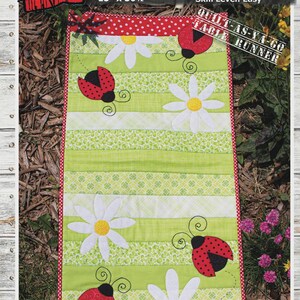 May include: A green and white table runner with ladybug and daisy appliqués. The runner is 15 inches by 30.5 inches and features the text "BUSY LADY BUGS". The skill level is easy.
