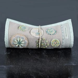May include: A handmade, rolled fabric item with embroidered floral designs in shades of blue, green, and beige. The item is secured with a beige string and features the text "one of a kind" on the side. The edges have a light blue patterned border.