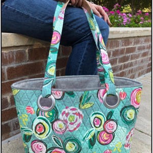 May include: A large tote bag with a floral pattern in shades of pink, yellow, and green on a teal background. The bag has a cork base, gray lining, and silver metal ring handles. The text "The Big Bag" is visible.