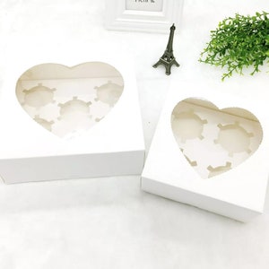 May include: Two white cardboard boxes with heart-shaped windows. The boxes are designed to hold cupcakes or other small treats. The boxes are empty.