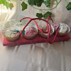 May include: A clear plastic box with a red handle contains four decorated cupcakes. The cupcakes are topped with pink, white, and green frosting and have gold and white accents. The cupcakes are sitting on a red base.