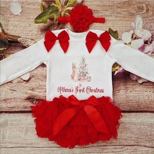 May include: A white long-sleeved onesie with red bows and a red tutu skirt. The onesie features a Christmas tree and bunny graphic, with the text "Athena's First Christmas". A red bow headband completes the outfit.