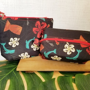 May include: Two black zippered pouches with a tropical print featuring red hibiscus flowers, white plumeria flowers, teal blue whales, and green leaves. The pouches have red zippers and the text "Aloha" is printed on the pouches.