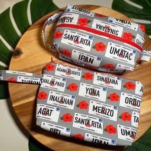 May include: Two grey zippered pouches with a floral print. The pouches feature a pattern of red hibiscus flowers and white license plates with the names of different towns and villages in Guam. The text on the license plates reads "Guam - Marianas" and "Tano I Chamoru".