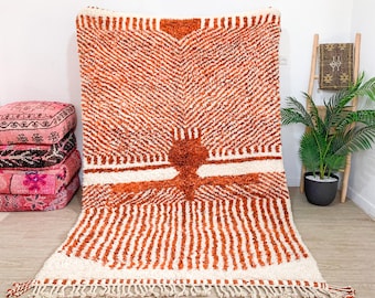 May include: A large, hand-woven rug with a geometric pattern in shades of white and orange. The rug has a fringe border and is made of wool.