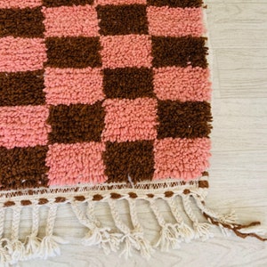 May include: A pink and brown checkered rug with a white fringe. The rug is made of a soft, fluffy material.