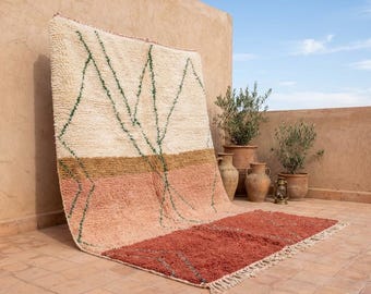 May include: A rectangular rug with a cream upper section, green geometric lines, a brown mid-section, and a reddish-orange lower section. The rug has a fringed edge and is displayed outdoors against a beige wall.