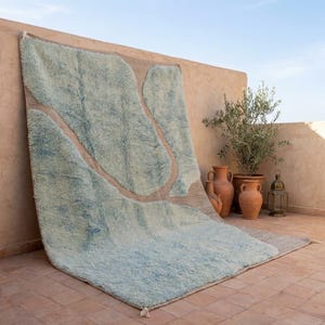 May include: A large, rectangular rug with a light blue, cloud-like design against a neutral background. The rug is displayed outdoors, leaning against a wall. Decorative pottery and plants are visible in the background.