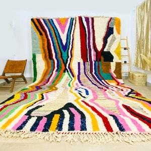 May include: A colorful Moroccan rug with a geometric pattern in shades of pink, yellow, blue, green, red, and white. The rug has a fringe border and is made of wool.