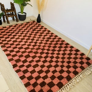 May include: A pink and brown checkered rug with fringe. The rug has a geometric pattern and is made of a soft, fluffy material.