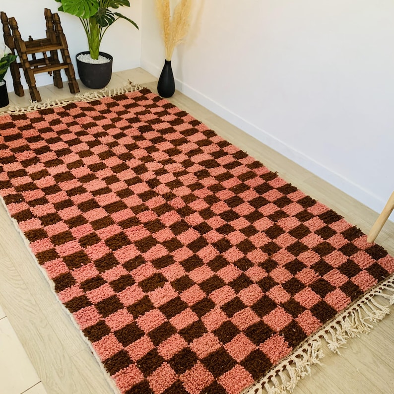 May include: A pink and brown checkered rug with fringe. The rug has a geometric pattern and is made of a soft, fluffy material.