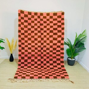 May include: A pink and brown checkered rug with a fringe border. The rug is made of a soft, fluffy material.